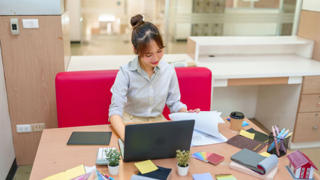 An Asian woman in her early career is working on a laptop at her desk, surrounded by blueprints, color swatches, and design materials. She is planning interior design concepts.の写真素材