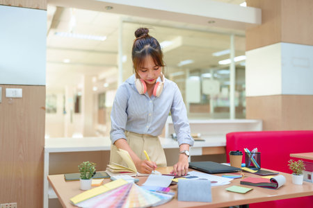 Asian woman interior designer at work, sketching ideas with color swatches and material samples. Modern office workspace with a creative young professional, design process for home decor and renovations.の写真素材