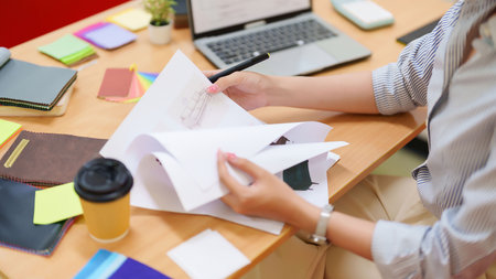 An adult Asian woman interior designer reviews blueprints, material swatches, and project details on a laptop while enjoying coffee. She is planning space designs and interior project with workspace.の写真素材