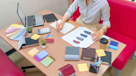 Adult Asian woman interior designer at her desk with color palettes, fabric swatches, furniture sketches, laptop and notepad. She is planning a home renovation project.の写真素材