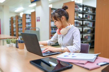 Young Asian woman studying in library, using laptop. Focus on education, research, online courses, student success. Bookshelves visible. Promote academic achievement and learning resources.の写真素材
