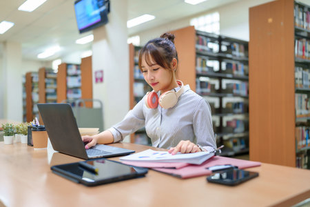 An Asian young woman focuses on her laptop at a library table, surrounded by bookshelves. She is studying, researching, and learning to ensure college university education for future career.の写真素材