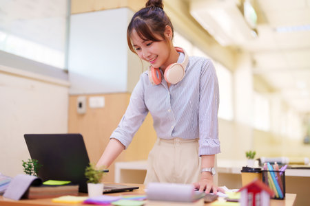 An Asian women studies online with a laptop and headphones on the desk with learning material. Education concept and smile in modern classroom for young adult.の写真素材