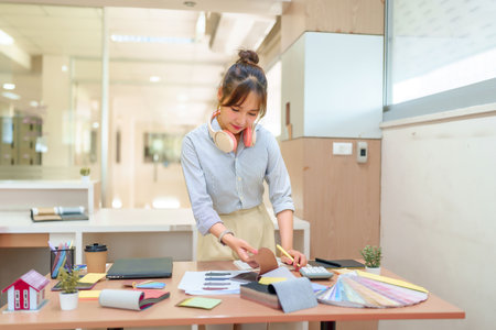 Asian young interior designer woman works with fabric samples and color palettes at her office desk. She selects materials for home renovation projects, crafting beautiful and functional spaces.の写真素材