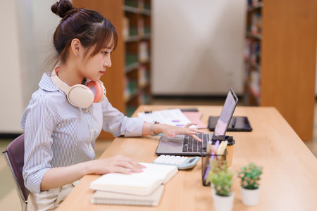 Young Asian woman using a laptop in a library. She is focused on her online studies and coursework, working on an academic project, and researching for her college assignments.の写真素材