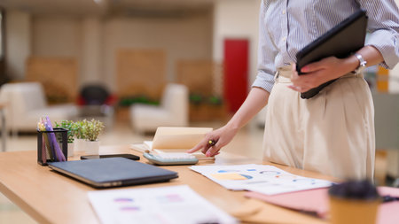 An Asian adult woman is using a calculator to analyze financial data charts in an office. This image is perfect for illustrating education, finance, or business-related content.の写真素材