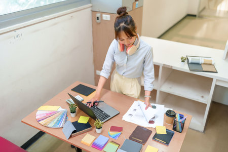 An Asian adult woman interior designer working at her desk with a laptop, color swatches, fabric samples, and design sketches. Brainstorming ideas for design projects.の写真素材