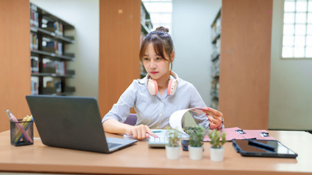 Asian young woman studying in a library with a laptop, calculator, and book. Preparing for academic research and exams, she embodies educational success. This image showcases dedication to learning.の写真素材