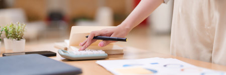 Close-up of an adult Asian woman calculating finances with a calculator, analyzing business charts at a desk. Concept of accounting, budgeting, financial planning in an educational setting.の写真素材