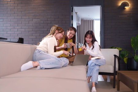 Three Asian young women enjoying online shopping together at home using laptop and credit card. Happy friendship moment relaxing during vacation holiday lifestyle.の写真素材