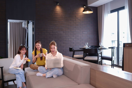 Three young Asian women are shopping online on a laptop while sitting on a sofa in their home. One woman is holding a credit card, and the others are browsing for products and services. Happy and relaxed lifestyleの写真素材