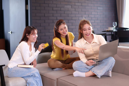 Three young Asian women enjoy shopping online using a laptop while relaxing on the sofa at home holding a credit card having fun and happy lifestyle with technologyの写真素材