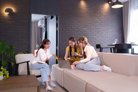 Three adult Asian women are happily shopping online using a laptop on the sofa at home. One woman is...