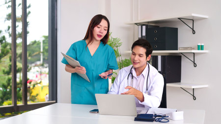 An Asian adult doctor is sitting at the desk with a laptop and discussing patient data with an Asian young female nurse holding a clipboard. They are reviewing medical records, collaborating in the clinic for a comprehensive healthcare examination treatment diagnosis.の写真素材