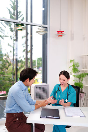 An Asian adult female doctor wearing scrubs uses a pen to examine an adult male patient's wrist and hands during a medical consultation in a clinic. They are seated at a table in an office.の写真素材