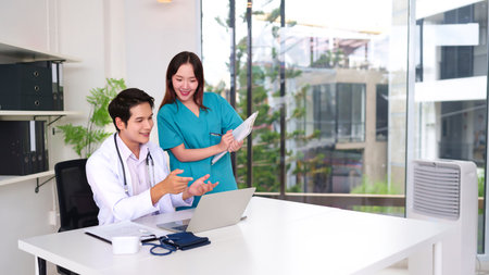 Young Asian doctor and nurse reviewing patient information on a laptop in a modern clinic. Healthcare professionals collaborating on treatment plans. Focus on teamwork and medical expertise in office setting.の写真素材