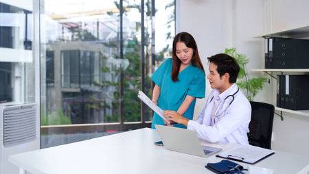 An Asian adult doctor and nurse are reviewing patient data together in a modern clinic. They are analyzing report for better healthcare. The medical professionals are working together in hospital.の写真素材