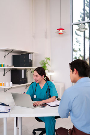 Young Asian women doctor is consulting with a patient, using a laptop to review information. The scene takes place in a well-lit clinic or modern hospital exam room.の写真素材