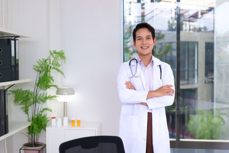 An adult Asian doctor stands with arms crossed, smiling confidently in a clinic setting. He wears a white coat and stethoscope, projecting a professional image for healthcare.の写真素材