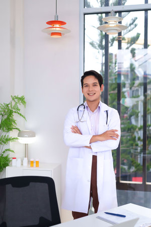 Portrait of a cheerful adult Asian male doctor wearing a white coat and stethoscope in a bright modern office ready to consult and help. Perfect for healthcare or medical related designs.の写真素材