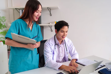 Adult Asian doctor is using a laptop for patient data with nurse holding a medical chart in clinic. They collaborate on patient care in a modern office setting.の写真素材