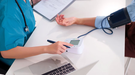 An Asian nurse in scrubs is using a digital blood pressure monitor to examine an adult patient's...