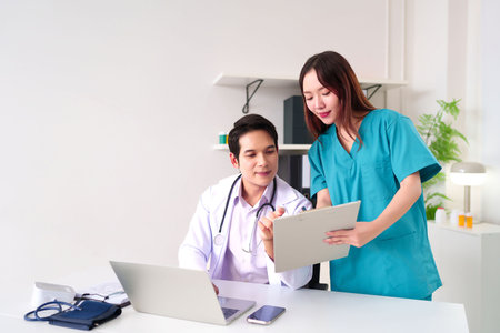 Asian adult doctor and nurse reviewing patient data on a clipboard using a laptop in a bright clinic setting. Ideal for healthcare, medicine, and professional teamwork related projects.の写真素材