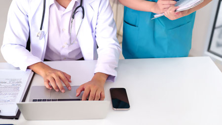 An Asian adult doctor is at a desk using a laptop in a clinic. The doctor is in a white coat, with a stethoscope around their neck and nurse taking notes. They're working with electronic patient records in a hospital office.の写真素材
