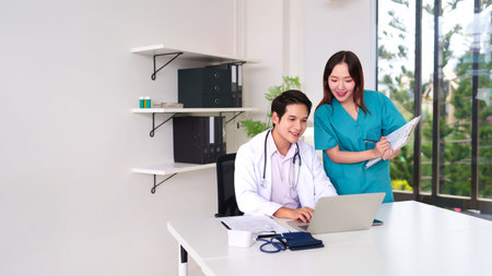 Asian adult doctor is working on laptop with nurse standing next to him discussing patient data in clinic office. Teamwork provides better healthcare and medical treatment serviceの写真素材