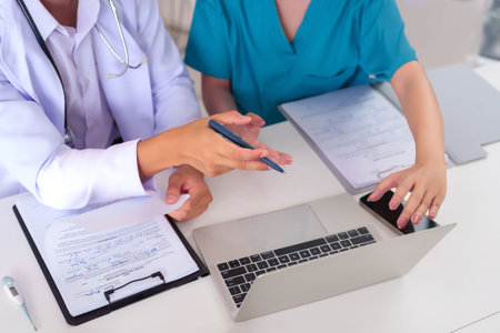 Asian adult doctors are collaborating on patient care reviewing documents on a laptop at their desk they are discussing health information treatment plans sharing insights for better diagnosis and careの写真素材