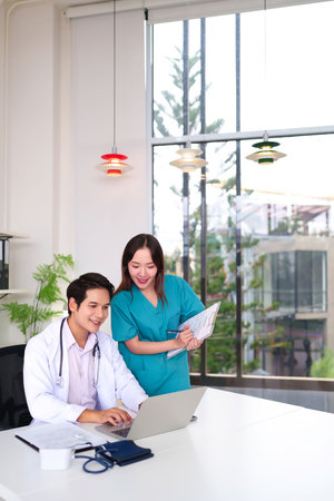 Young Asian adult doctor using laptop with his colleague nurse is holding document of medical record in bright modern clinic. Healthcare teamwork concept.の写真素材