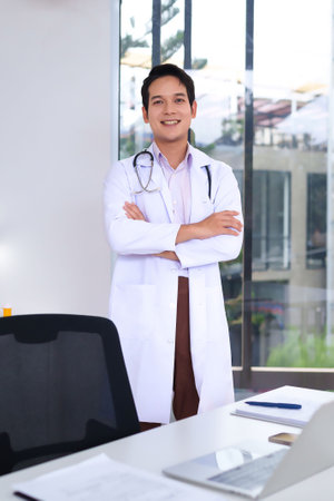 Smiling Asian adult doctor standing in office with stethoscope expertise. He is wearing a white coat. Professional healthcare expert at workplace in clinic. Patient care consultationの写真素材