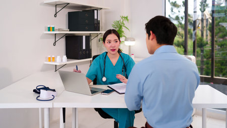 Asian adult woman doctor in scrubs consulting with a male patient in a clinic exam room. They are discussing healthcare advice and medical treatment planning in the hospital office.の写真素材
