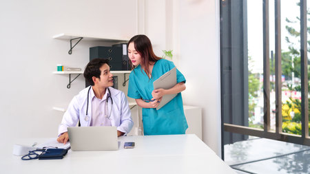 An Asian adult doctor and nurse are discussing a patient case while sitting at a desk using a laptop. This image shows teamwork between medical professionals in a bright clinic.の写真素材