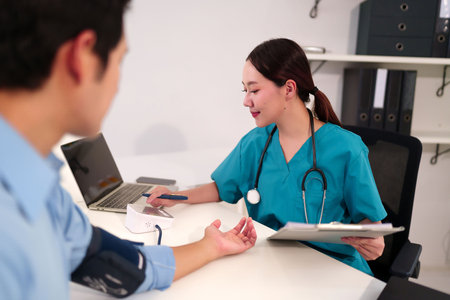 Asian woman doctor in green scrubs checks blood pressure of adult patient using digital monitor during medical consultation writes report healthcare in clinic.の写真素材