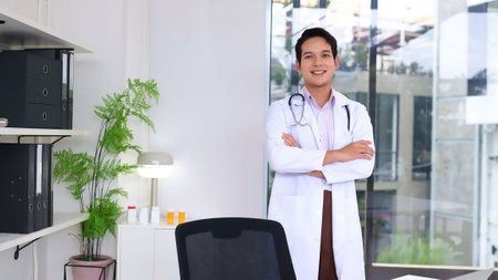 A confident adult Asian doctor stands in their office. They are smiling wearing a white coat and a stethoscope. It is a professional setting for medical healthcare work.の写真素材