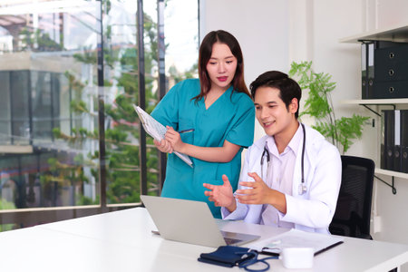 Two young adult Asian medical professionals doctor and nurse collaborating on patient care using a laptop in a bright modern clinic fostering teamwork and healthcare excellenceの写真素材