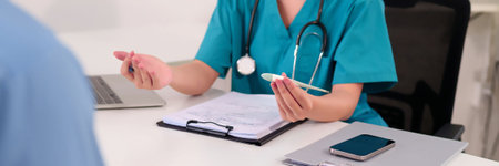 An Asian adult female doctor wearing a stethoscope holds a thermometer while consulting with a patient in a clinic office explaining diagnosis and treatment options.の写真素材