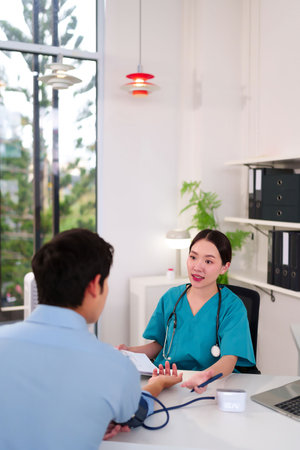 An Asian female physician checks the blood pressure of an adult male during a medical consultation. They discuss health in a modern clinic setting for better treatmentの写真素材