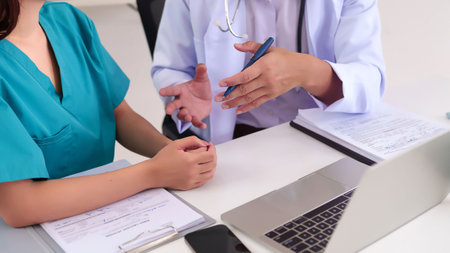 Adult Asian doctor explaining diagnosis to Asian nurse with laptop and medical report during healthcare discussion in a bright hospital office providing expert advice.の写真素材