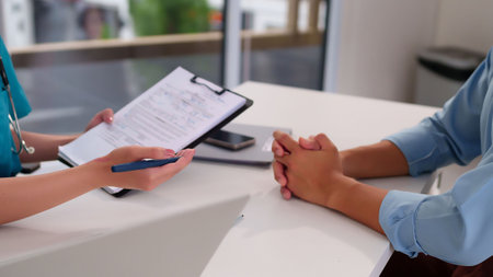 An Asian woman doctor's hands holds a pen and clipboard, explaining diagnosis to a patient in a clinic. The adult patient listens attentively receiving treatment and medical advice during examination.の写真素材