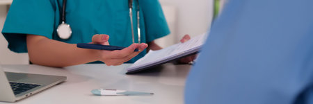 Asian adult female nurse in green scrubs reviews a medical chart with a pen in her hand during a patient consultation in clinic.  She analyzes the diagnosis with a laptop thermometerの写真素材
