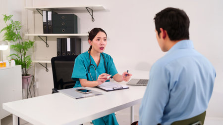 Asian female doctor in green scrubs explains diagnosis holding pen and thermometer to patient in a clinic office. Healthcare professional consultation medical exam and well being.の写真素材