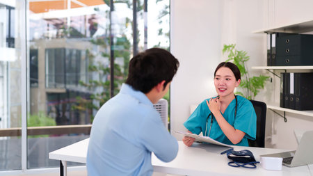 Adult Asian woman doctor consults adult man patient in clinic examination room for diagnosis. Professional healthcare setting with modern equipment for patient checkup and well being careの写真素材