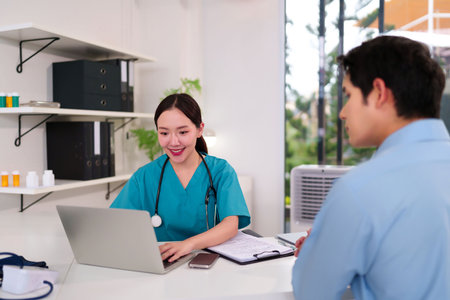 Young Asian female doctor in scrubs using a laptop to talk to a patient in a clinic setting. This captures the healthcare professional providing checkup examination with happy expression.の写真素材
