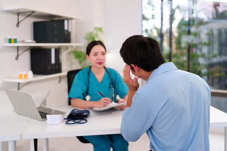 Asian adult female doctor in green uniform consults with male patient who is experiencing a headache in the clinic office. The doctor is taking notes while the man is sick healthcareの写真素材