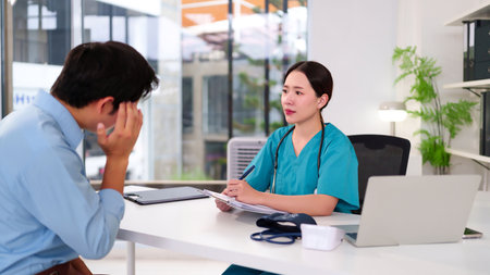 An Asian female doctor in scrubs consults a young adult male patient about his health problems offering medical advice and diagnosis in a modern clinic setting promoting wellness.の写真素材