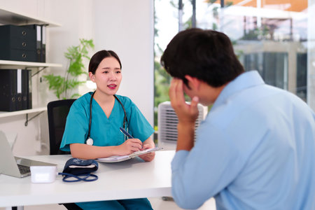 An Asian young adult nurse in scrubs consults with an adult male patient in a clinic, taking notes. She explains his diagnosis in a calm and reassuring manner in office setting.の写真素材
