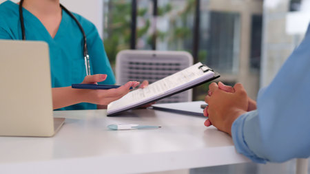 An Asian adult female doctor in scrubs discusses diagnosis and treatment plan with patient in clinic office. Medical professional consulting healthcare with thermometer on table.の写真素材