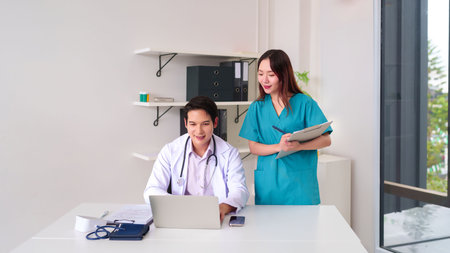 Asian adult doctor uses laptop at desk with nurse Asian adult reviewing patient chart in clinic. Medical professionals collaborating on healthcare. Patient consultation for better careの写真素材